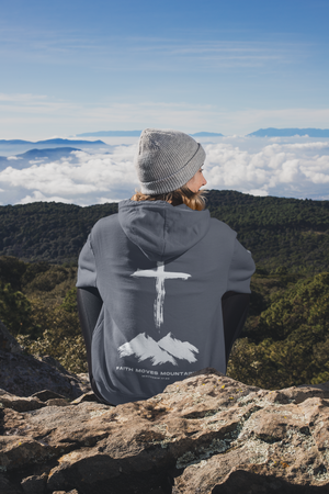 Person sitting on mountain peak wearing Faith Moves Mountains hoodie in gray with white cross and mountain graphic