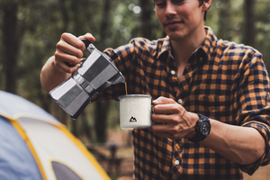 Man pouring coffee into a Mountain Faith Co. camp mug in a forest setting, outdoor faith lifestyle image.