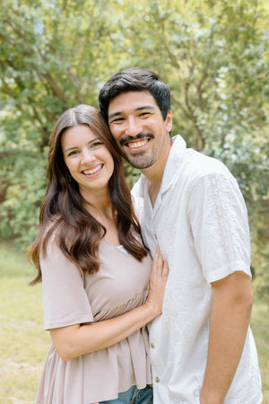 Smiling couple standing together outdoors in a bright forest setting, warm lifestyle portrait.